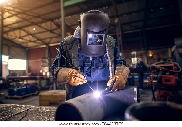 Close up portrait view of professional mask
protected welder man in uniform working on the metal sculpture at
the table in the industrial fabric workshop in front of few other
workers.