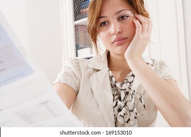 Close Up Portrait View Of A Young Attractive Professional Business Woman Sitting At Her Working Desk And Looking At A Bank Statement Bill, Thoughtful And Worried. Office Interior.