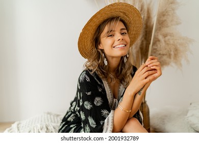 Close up portrait of woman in bohemian dress sitting on pillows at home. Wearing earrings with feather  and bracelets. White background in studio with pampas grass decor.