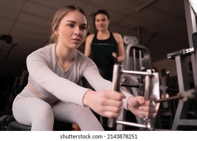 Close Up Portrait Of A Woman In A Health Club With Her Personal Trainer Learning Proper Technique On A Weight Lifting Machine Pulling Machine In A Fitness Club.