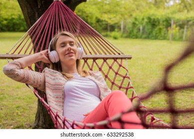 Close Up Portrait Of Woman Lying Down On Hammock Listening To Music With Cell Phone. Cheerful Girl Enjoy In Red Hammock Outdoor. Woman Relaxing Outside Listening To Music With Earphones