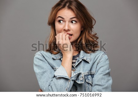 Close up portrait of a worried teenage girl in denim jacket biting her nails and looking away isolated over gray background