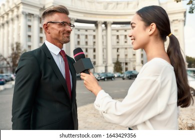 Close Up Portrait Of Young Asian Woman Conducting Journalistic Interview Of Cheerful Politician. People Making Interview Using Equipment Set At Outdoor Location. Side View. Horizontal Shot