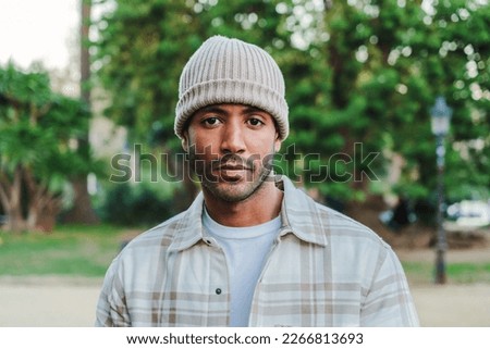 Close up portrait of young attractive african american man with beanie hat looking serious at camera. Front view of a hispanic guy standing in a park outdoors with sad attitude. High quality photo