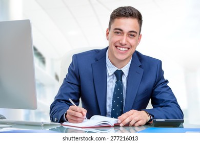 Close Up Portrait Of Young Attractive Businessman In Blue Suit At Desk In Office. Young Man Writing In Agenda With Pen.