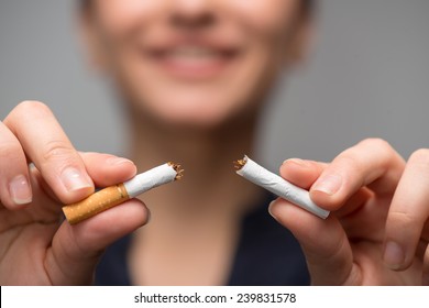 Close up portrait of young attractive woman breaking down cigarette to pieces. Studio shot selective focus isolated on grey. Addiction concept