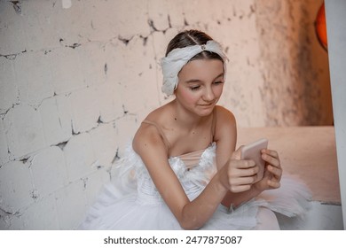 Close up portrait of young ballerina in white tutu, feather tiara, sits on the steps in loft interior, holds smartphone in hands, surfs the Internet. Teenager social media. Break between rehearsals - Powered by Shutterstock