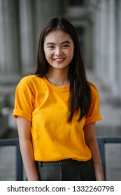 Close up portrait of a young, beautiful, delicate featured elfin Chinese Asian girl model. She is wearing a yellow tee and standing against a grey background of courtroom pillars.  She is smiling. 