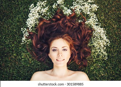 Close Up Portrait Of Young Beautiful Girl Woman With Red Brown Hair Lying On Grass With White Small Flowers Around Her Head .View From Above Top Overhead. Concept Of Spring Summer Youth Happiness