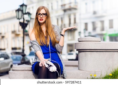 Close up portrait of young  blonde woman with sad and wailful emotional face , holding earphones, sitting on sidewalk , old european city background. Wearing blue dress and trendy silver handbag. 