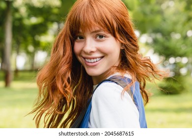 Close up portrait of a young caucasian woman girl teenager student schoolgirl with ginger red hair and toothy smile walking in park forest outdoors looking at camera.