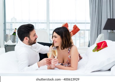 Close Up Portrait Of Young Couple On Honeymoon In Hotel.Man And Woman Drinking Champagne On Bed In Suite.