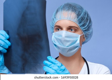 Close Up Portrait Of Young Doctor Holding X-ray Roentgen In Hands And Making Analysis, Female Wearing Disposable Hat, Rubber Gloves, Protective Medical Mask, Treating Patient With Serious Disease.