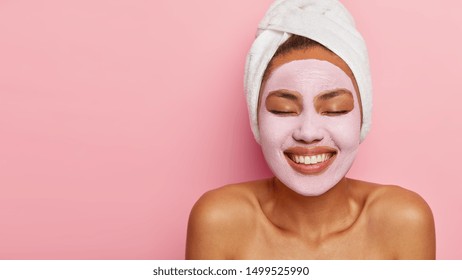 Close Up Portrait Of Young Female Model Applies Homemade Facial Clay Mask, Has White Towel Wrapped Around Head, Keeps Eyes Shut, Smiles Happily, Models Against Pink Background. Beauty Treatment