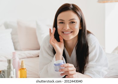 Close up portrait of a young girl woman using softening moisturizing face cream for skin face care. Beauty treatment. Woman applying cosmetics on her face relaxing in spa bathrobe on her bed