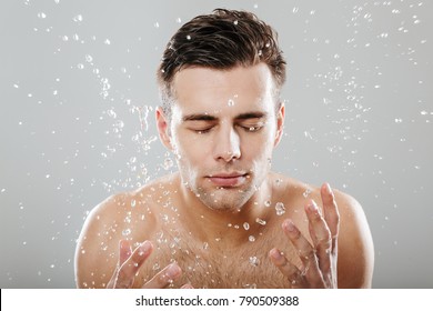 Close up portrait of a young half naked man surrounded by water drops washing his face isolated over gray background