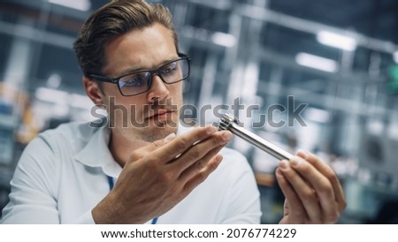 Close Up Portrait of Young Handsome Engineer in Glasses Working on Manufacturing Metal Parts in Office at Car Assembly Plant. Industrial Product Designer Examining Prototype Parts Before Production.