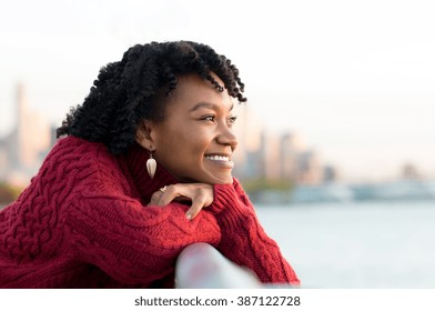 Close up portrait of a young happy african woman leaning on the banister of a bridge near river. Happy young african woman at river side thinking. Smiling pensive girl looking across river at sunset.
