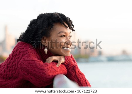 Close up portrait of a young happy african woman leaning on the banister of a bridge near river. Happy young african woman at river side thinking. Smiling pensive girl looking across river at sunset.
