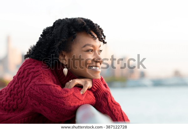 Close up portrait of a young happy african\
woman leaning on the banister of a bridge near river. Happy young\
african woman at river side thinking. Smiling pensive girl looking\
across river at sunset.\

