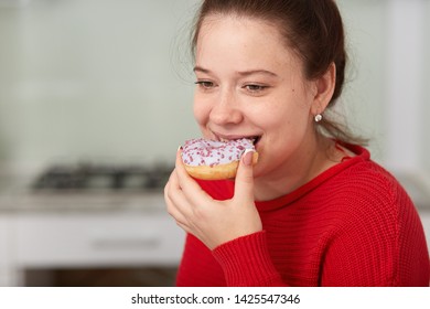 Close Up Portrait Of Young Happy Woman Eating Cake While Sitting At Table In Kitchen, Lady Wearing Red Casual Sweater, Has Happy Facial Expression, Looking Down. Unhealthy Eating And Lifestyle Concept