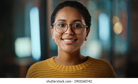 Close Up Portrait Of A Young Latina With Short Dark Hair And Glasses Posing For Camera In Creative Office. Beautiful Diverse Multiethnic Hispanic Female Wearing Yellow Jumper Is Happy And Smiling.