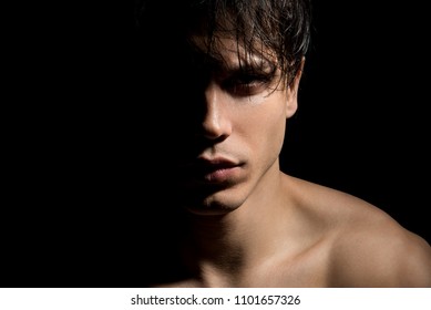Close up portrait of young mysterious guy with half face in shadow. He is standing on dark background and expressing thoughtfulness. Masculine beauty concept