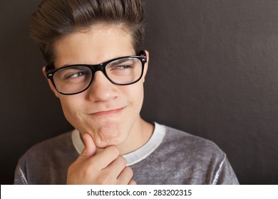 Close Up Portrait Of A Young Naughty Teenager Man With A Mischievous Thoughtful Expression, Looking Away Against A Dark Background, Wearing Reading Glasses, Home Interior. Quirky Young Man Indoors.