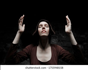 Close up portrait of young nun. Nun is praying. Photo on black dark background. closed eyes. arms raised to the heaven.