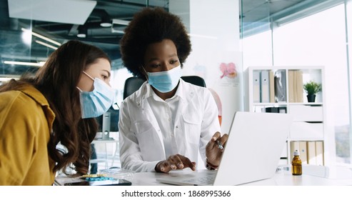 Close Up Portrait Of Young Pretty African American Woman Doctor In Medical Mask Sit At Workplace Speaking On Consultation With Female Patient. Coronavirus Covid-19. Medical Center Concept