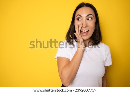 A close up portrait of a young pretty woman telling secret information, holding her hand near mouth and standing against yellow background