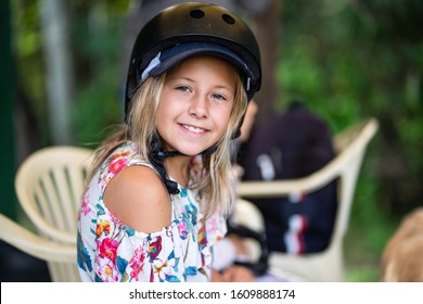 A Close Up Portrait Of A Young Smiling Girl Wearing A Summer Top And A Horse Riding Helmet Sitting On A Chair