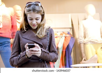 Close up portrait of a young stylish teenager woman using a smartphone device while visiting the city, leaning on a fashion store shop window while shopping for clothes.