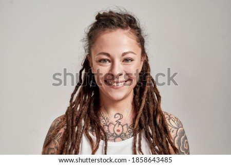 Close up portrait of young tattooed caucasian woman with dreadlocks wearing white shirt, smiling at camera while posing isolated over grey background. Front view. Horizontal shot