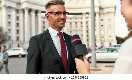 Close Up Portrait Of Young Woman Conducting Journalistic Interview Of Cheerful Politician. People Making Interview Using Equipment Set At Outdoor Location. Horizontal Shot. Selective Focus On Man