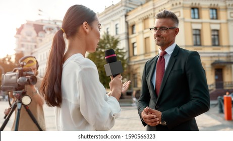 Close Up Portrait Of Young Woman Conducting Journalistic Interview Of Cheerful Politician. People Making Interview Using Equipment Set At Outdoor Location. Horizontal Shot