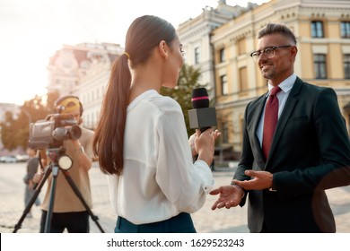 Close Up Portrait Of Young Woman Conducting Journalistic Interview Of Cheerful Politician. People Making Interview Using Equipment Set At Outdoor Location. Horizontal Shot. Low Angle