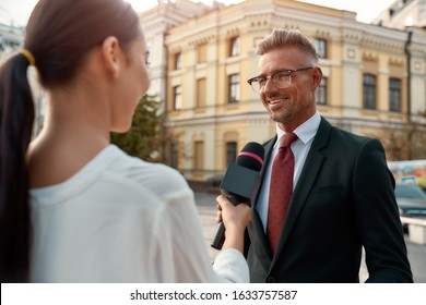 Close Up Portrait Of Young Woman Conducting Journalistic Interview Of Cheerful Politician. People Making Interview Using Equipment Set At Outdoor Location. Horizontal Shot. Selective Focus On Man