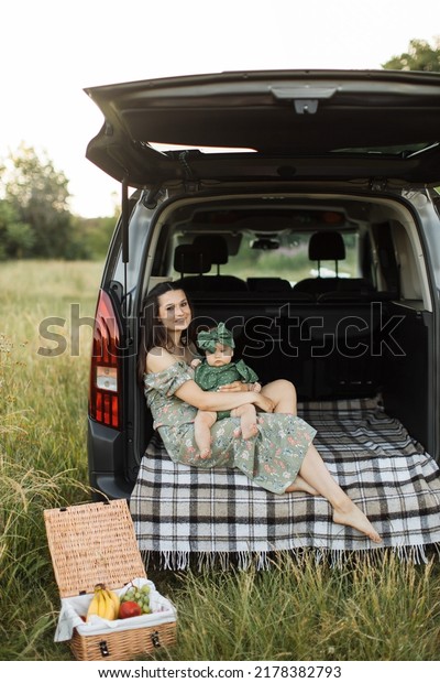 Close up portrait of young woman smiling and\
looking at camera while sitting on car trunk during picnic with her\
daughter on hands. Mother enjoying every moment spending with her\
loving child.