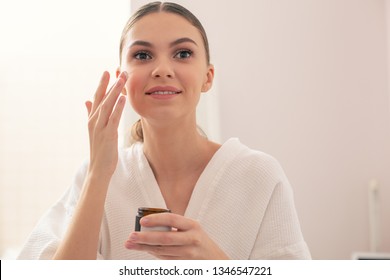 Close up of positive young woman in a bathrobe standing with a jar of cream in her hand and smiling while putting small amount of cream on her face - Shutterstock ID 1346547221