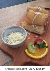 A Close Up Potrait Of Fried Cassava With Shredded Cheese On Bowl And Fruit Garnish. Served On Wood Board