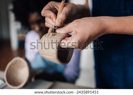 Close up of pottery class tutor standing at studio with clay work in hands and decorating pottery while showing technique to her students. Cropped picture of pottery course teacher decorating pottery.