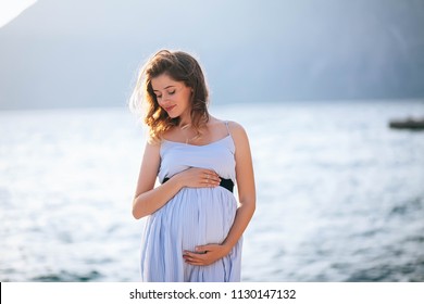 Close Up Pregnant Woman Hands Touching On Her Belly With Blurred Sea Background. Expectant Mother Wear White Maternity Dress In Summer Vacation For Relax. Pregnancy Healthy, Love And Travel Concept.