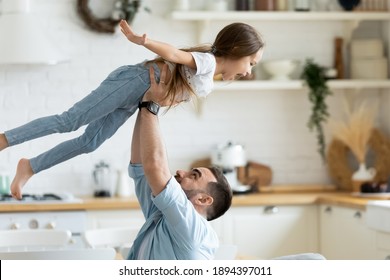 Close up preschool daughter flying with hands outstretched in caring father arms in modern kitchen. Happy smiling dad holding lifting cute girl. Family enjoying weekend at home together.