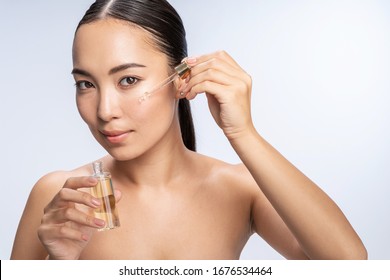 Close Up Of Pretty Asian Lady Applying Cosmetic Oil On Her Face With Pipette. Beauty Therapy Concept