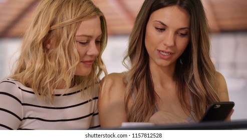 Close up of pretty brunette woman showing blonde friend smart phone inside house