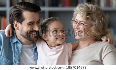 Close up pretty little girl hugging smiling young father and mature grandmother wearing glasses, family portrait cute preschool daughter granddaughter between loving dad and grandma at home