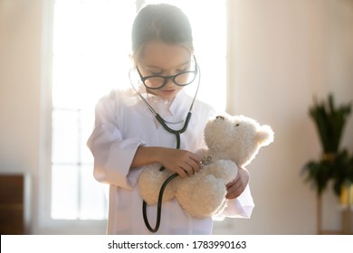 Close Up Pretty Little Girl Playing Doctor, Using Stethoscope, Holding Fluffy Toy Bear, Checking Heart Or Breath, Preschool Child Wearing White Coat Uniform And Glasses Pretending Pediatrician