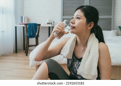 Close Up Pretty Sweaty Japanese Lady With A Towel Around Neck Is Drinking A Glass Of Water After Morning Training In A Bright Bedroom At Home.