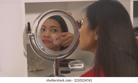 CLOSE UP Pretty Woman Self Massaging Her Face With Jade Scraper For Rejuvenation. Reflection Of Beautiful Asian Woman In Cosmetic Mirror Performing Anti-aging Massage Treatment With Gua Sha Scraper.